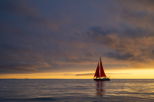 red sailboat sailing among in Disko Bay glacier with orange sunset during polar summer midnight solar season. Ilulissat, Greenland