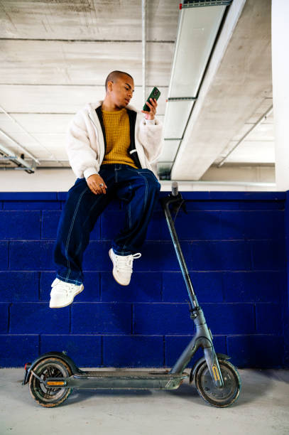 istock Young man using mobile phone while sitting on a wall with his electric scooter 2194822593