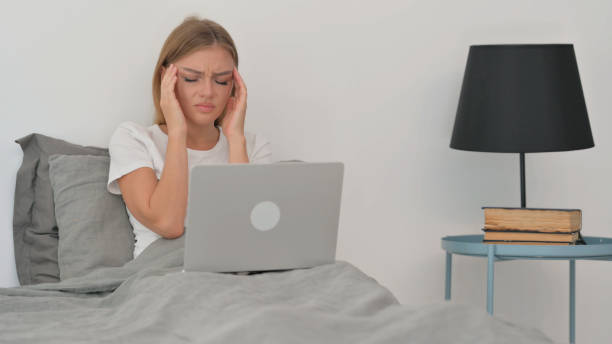 istock Young Woman with Headache Working on Laptop while Sitting in Bed 2194823997