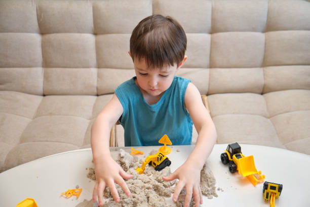 istock Child playing with kinetic sand and toy construction vehicles on a white table. Indoor activity. Front-facing portrait photography. Kid aged three year 2194845673