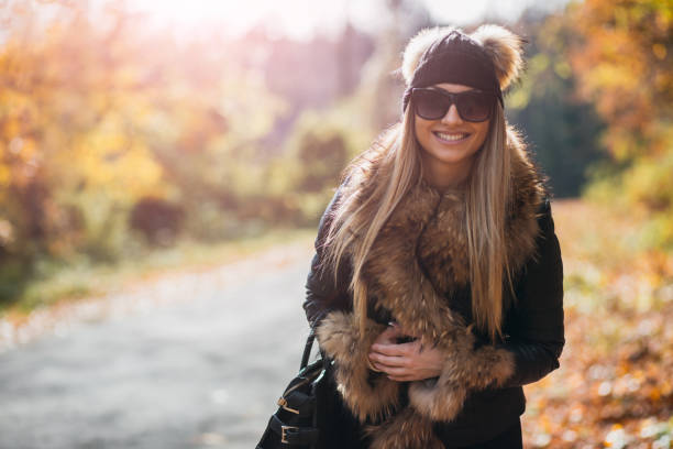A cheerful woman dressed warmly in stylish attire enjoys a sunny autumn day outdoors, surrounded by vibrant seasonal foliage. Her fashionable outfit highlights the cozy essence of fall trends