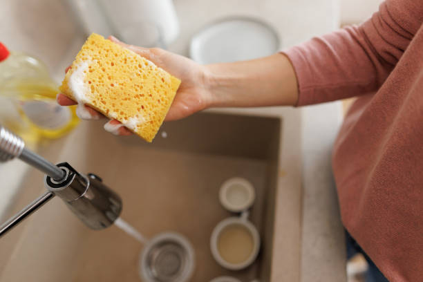 Woman holding a soapy sponge above a kitchen sink, preparing for dishwashing. Dish soap and dirty cups clutter the background, highlighting daily cleaning routines in a home setting