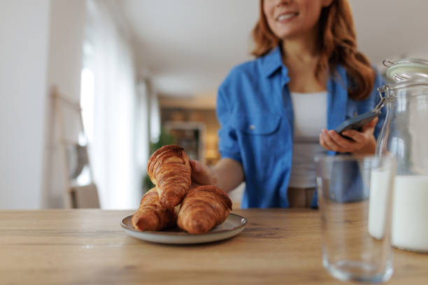 Woman placing croissants on table for breakfast while using phone