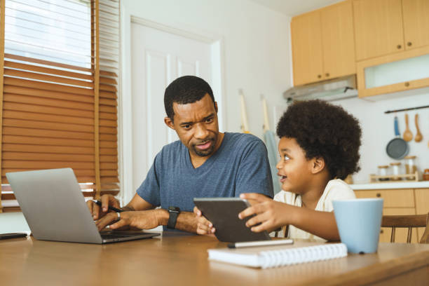 istock Black African American Father working on laptop while son uses tablet beside him at kitchen table, depicting focused and interactive family time. 2194873696