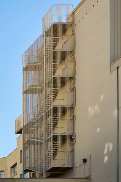 Exterior metal fire escape attached to the side of a beige building. The structure is enclosed with vertical metal bars and extends over several floors, reflecting urban safety features.