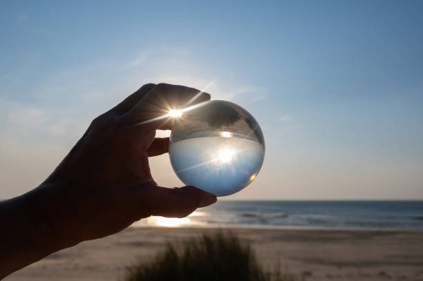 A hand of an  person holds a glass ball, the sea and the setting sun are reflected in the ball