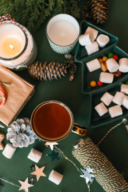 istock Cozy Christmas setup featuring a wrapped gift with a candy cane, a lit candle, tea, an open book, pinecones, marshmallows, and evergreen branches on a green background 2194886902