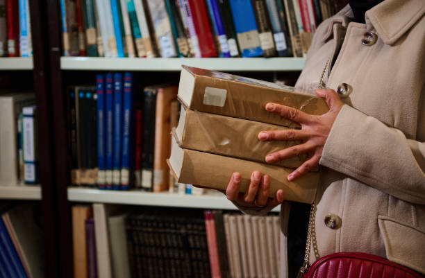 Close-up of a person grasping multiple books in a library, featuring bookshelves filled with a variety of literature. Highlights academic, educational, or literary contexts.