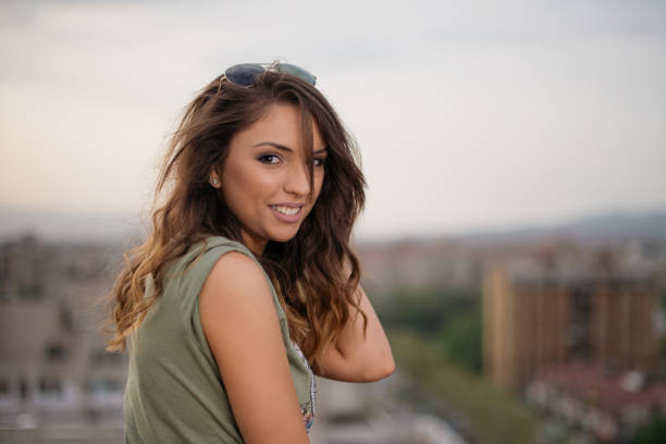A cheerful woman with wavy hair and sunglasses smiles while standing on a rooftop overlooking a cityscape