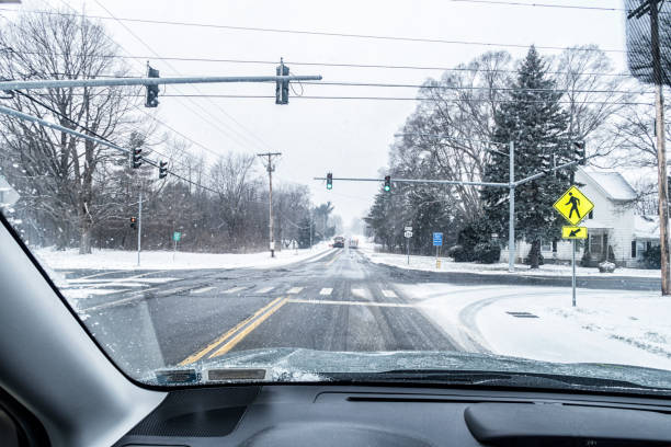 Car driver POV looking through windshield across a traffic signal road intersection at a large municipal snowplow dump truck approaching from the opposite direction just as it passes an ambulance rushing ahead with its emergency lights flashing during a January winter blizzard snow storm on a sloppy, slippery, rural highway near Rochester, in western New York State.