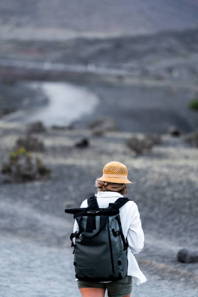 Woman traveler walks on a rocky trail wearing a hat and carrying a backpack, surrounded by barren scenery.