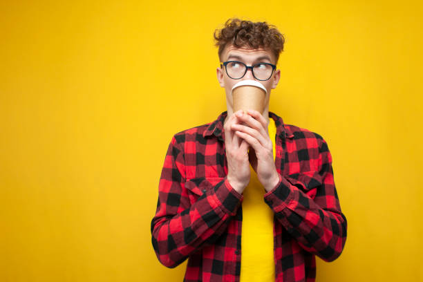 istock curly guy in glasses drinks coffee on a yellow background, a student holds coffee and looks at copy space 2194954555
