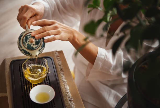 Female hands pouring tea from patterned clay teapot into clear glass pitcher, creating serene tea ritual. Soft light, wooden tea tray and greenery evoke calming mindful chenese ceremony atmosphere. manos femeninas vertiendo té de una tetera de arcilla estampada en una jarra de vidrio transparente, creando un ritual de té sereno. la luz suave, la bandeja de té de madera y la vegetación evocan un ambiente relajante y consciente de la ceremonia chen