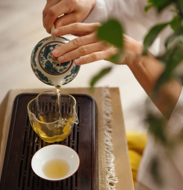 istock Female hands pouring tea from patterned clay teapot into clear glass pitcher, creating serene tea ritual. Soft light, wooden tea tray and greenery evoke calming mindful chenese ceremony atmosphere. 2194979223