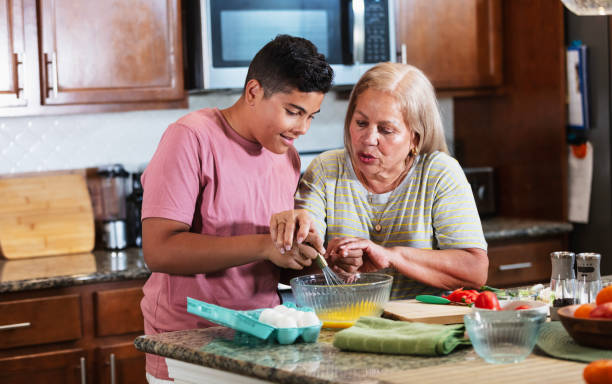 A senior Hispanic woman, in her 70s, in the kitchen, cooking with her 12 year old grandson. Grandma is trying to show the boy how to properly whisk eggs in a mixing bowl.