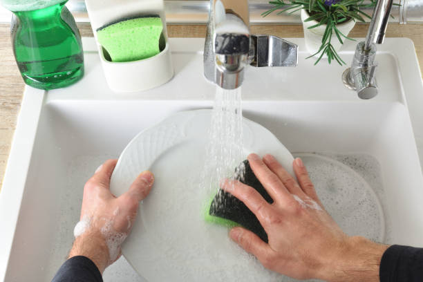 Detail of hands washing dishes in a sink with tap running water and cleaning tools around stock photo