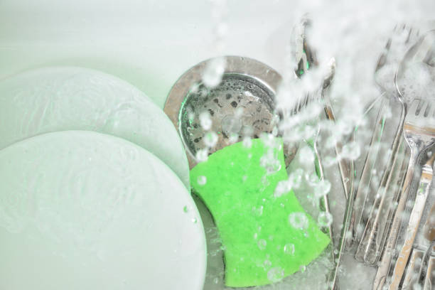 White sink with white dishes covered in metal and green scouring pad with water dripping on it. Top view.