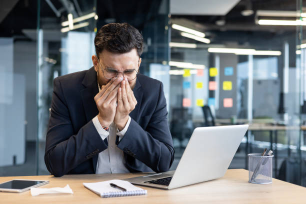 istock Sick man at workplace, has runny nose sneezing, allergic attack. Businessman inside office working with laptop 2195050120