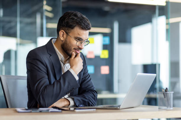 istock Thinking businessman at workplace inside office, man in business suit working with laptop, reading online report, and preparing presentation investment project 2195050414