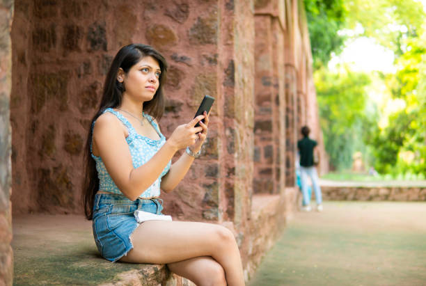 Tourist young woman looking away and contemplating while using her smartphone at sitting on an ancient brick wall at Humayun’s Tomb, the famous landmark and enjoying her leisure time.