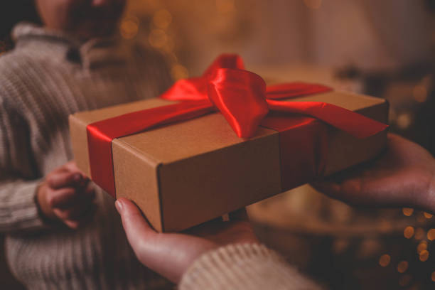 istock Hands exchanging a festive gift box with a red ribbon under a beautifully decorated Christmas tree. Cozy holiday setting with candles, miniature houses, and a warm atmosphere 2195051453