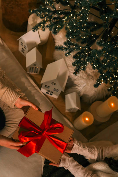 istock Hands exchanging a festive gift box with a red ribbon under a beautifully decorated Christmas tree. Cozy holiday setting with candles, miniature houses, and a warm atmosphere 2195051697