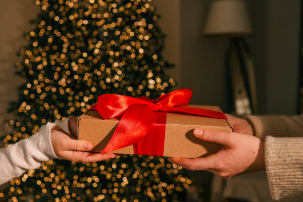 istock Hands exchanging a festive gift box with a red ribbon under a beautifully decorated Christmas tree. Cozy holiday setting with candles, miniature houses, and a warm atmosphere 2195051709