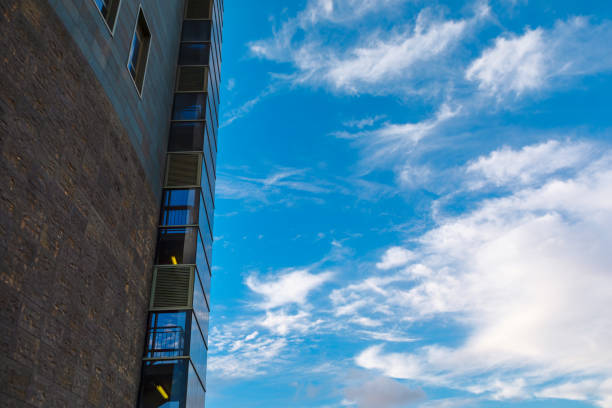Modern building facade with a mix of brick and glass panels stands against a vibrant blue sky filled with scattered white clouds, creating a striking contrast of textures and colors