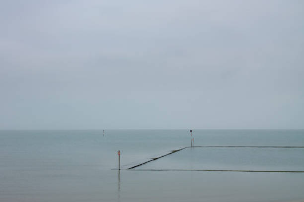 A calm sea showing a horizon and muted colours in the sky and water, with a almost submerged tide pool and posts.