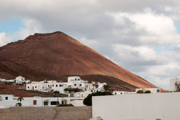 Village with traditional simple houses with white facade. High mountain of a red color in the background. Cloudy sky in the winter. Soo, Lanzarote, Canary Islands, Spain.