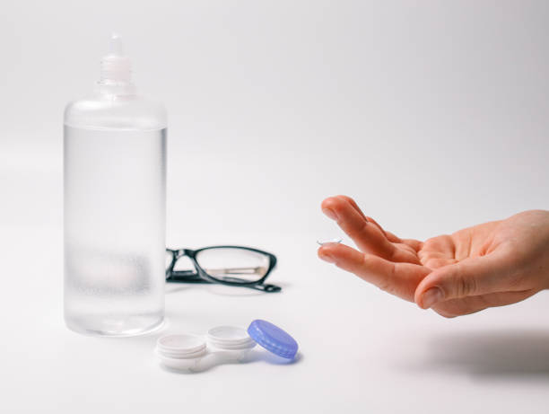Hand holding a transparent contact lens next to lens case, solution bottle, and glasses on a white background. Concept of eye care, vision correction, and optical hygiene