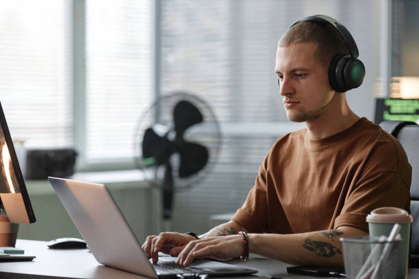 istock Focused Male Programmer in Headphones Using Laptop Working Indoors 2195099697