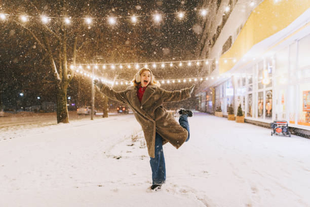 A woman with long hair in a warm coat and knit hat spins with open arms, enjoying the snowfall at a holiday market filled with twinkling lights and decorations.