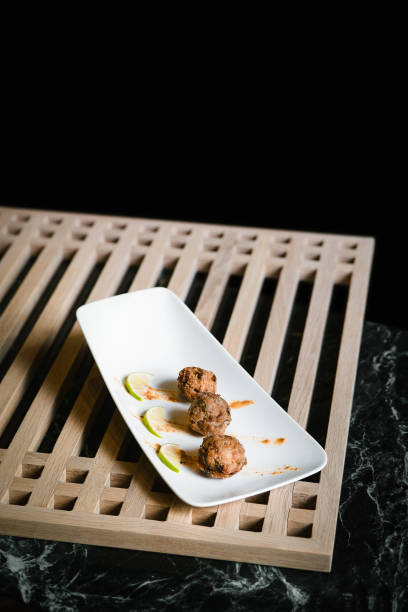 This image features a fusion-style Thai appetizer of meatballs, elegantly drizzled with a flavorful savory sauce. The dish is garnished with lime wedges, adding a touch of freshness and color contrast. Presented on a sleek rectangular white plate atop a s