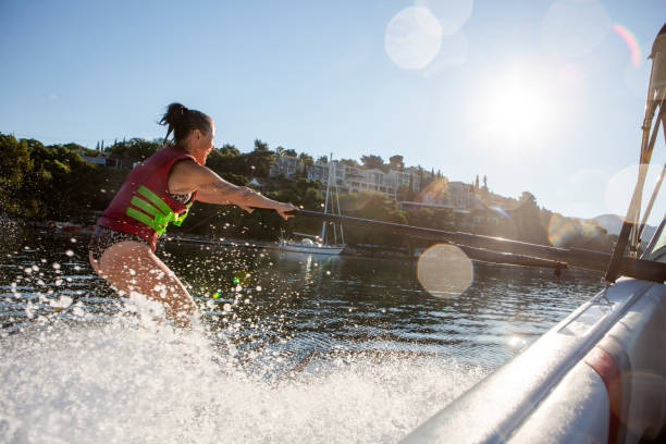 Young beautiful active blond woman riding on the wakesurf holding rope of motorboat on the background of trees