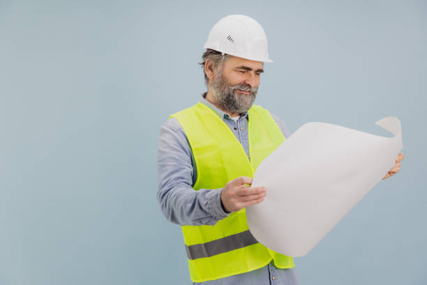 istock Senior engineer wearing safety helmet and vest reading blueprints on blue background 2195135033