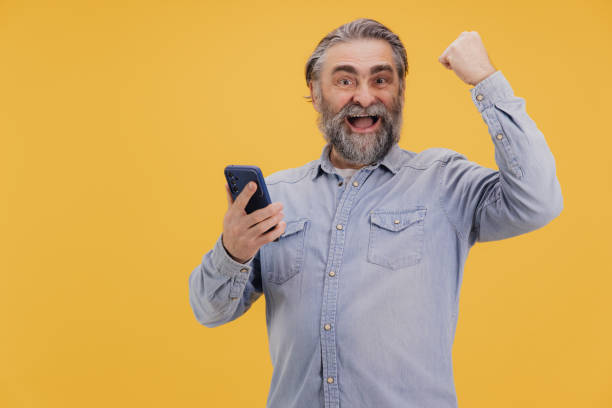 istock Excited senior man celebrating success on smartphone on yellow background 2195135302