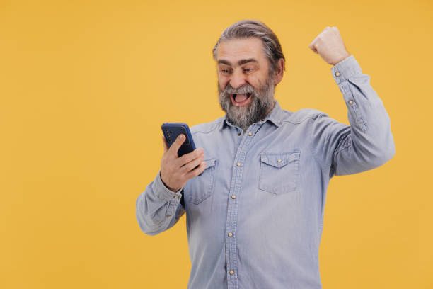istock Excited senior man celebrating success while looking at smartphone on yellow background 2195135323