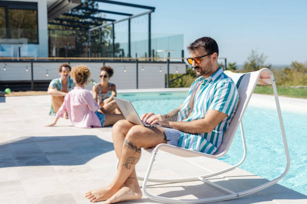A man works on his laptop while relaxing by the pool, enjoying a sunny day. His friends chat and unwind on the poolside, creating a perfect blend of productivity and leisure in a vibrant summer setting.