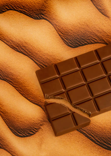 A milk chocolate bar with a broken piece placed on wavy desert sand, creating a striking visual contrast between the creamy cocoa texture and the smooth, flowing dunes. The image captures the warmth and richness of chocolate in a conceptual gourmet food photography setting, emphasizing indulgence, natural beauty, and the interplay of textures.