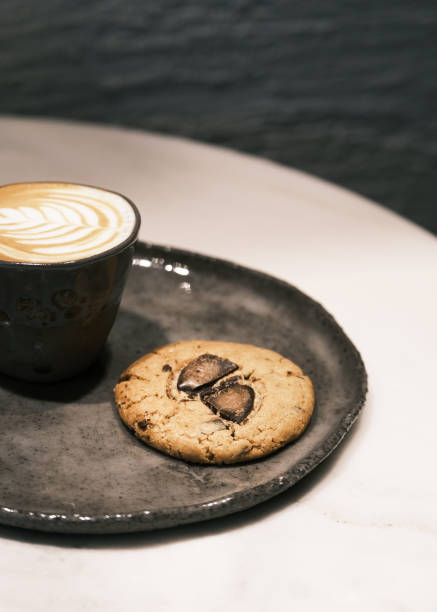 istock Artisan chocolate chip cookie with a gooey center on a rustic ceramic plate, served with a cup of creamy latte featuring intricate latte art, cozy café ambiance, handcrafted dessert concept, perfect for bakery branding and coffee shop aesthetics 2195180450