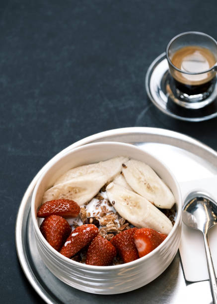 Top view of a nutritious breakfast bowl with granola, fresh strawberries, banana slices, and nuts, served on a silver tray with a spoon and espresso, minimalist dark background, healthy lifestyle concept