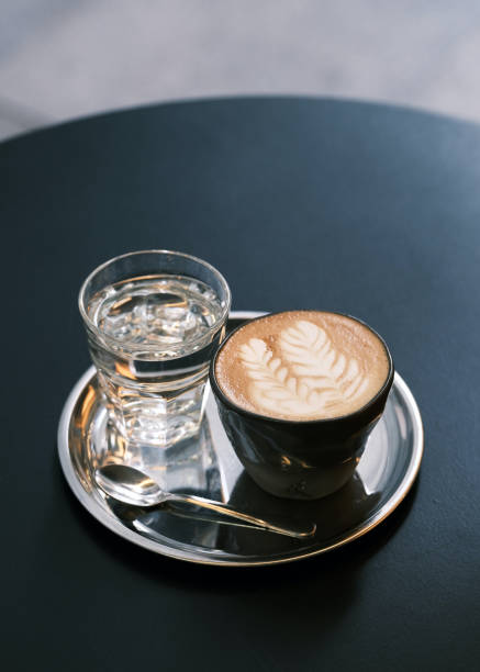Elegant coffee presentation with latte art in a glass cup on a silver tray, accompanied by a glass of water and spoon, modern café ambiance, specialty coffee culture, barista craftsmanship, minimalist coffee setup