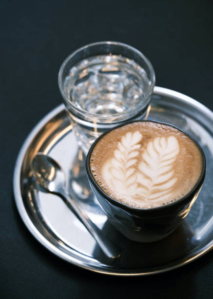 istock Elegant coffee presentation with latte art in a glass cup on a silver tray, accompanied by a glass of water and spoon, modern café ambiance, specialty coffee culture, barista craftsmanship, minimalist coffee setup 2195180541