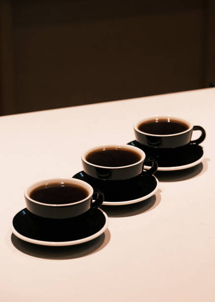 Three black ceramic coffee cups filled with freshly brewed coffee on a minimalist white table, warm lighting creating a cozy ambiance, specialty coffee tasting and modern café aesthetic concept