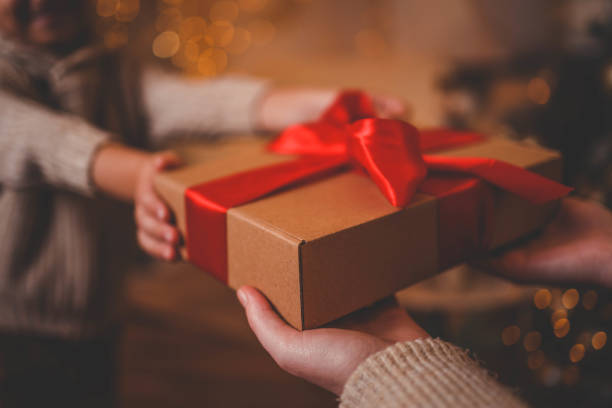 istock Hands exchanging a festive gift box with a red ribbon under a beautifully decorated Christmas tree. Cozy holiday setting with candles, miniature houses, and a warm atmosphere 2195212040
