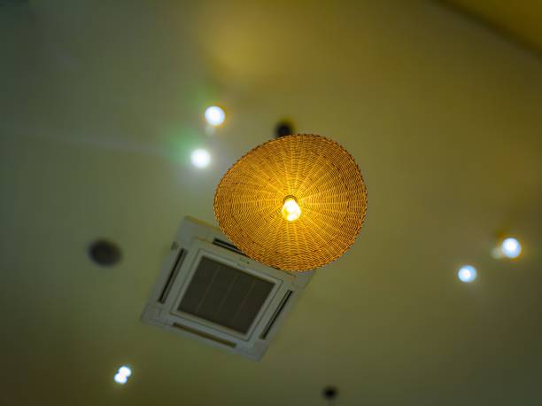 A warm, woven rattan lampshade with a glowing bulb hangs from a ceiling, surrounded by recessed lights and an air conditioning vent, creating a cozy ambiance