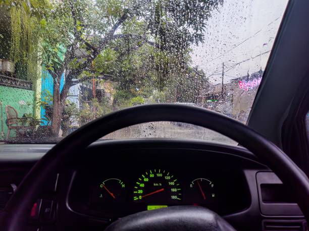 A view from inside a car during a rainy day, showing the wet windshield with raindrops, the steering wheel, and the dashboard with illuminated gauges