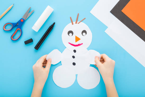 istock Little child boy hands holding and creating snowman shape from white paper on light blue table background. Pastel color. Making winter elements. Closeup. Point of view shot. Top down view. 2195227830