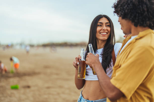Two friends sharing a joyful moment at the beach while holding drinks, with sandy shores and a calm atmosphere creating a lively and cheerful environment.